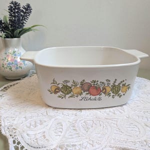 May include: White rectangular casserole dish with handles, decorated with a colourful illustration of various vegetables and the text "L'échalote". The dish is on a white lace doily.