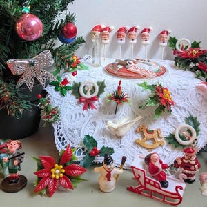 May include: A collection of vintage Christmas ornaments and figurines including Santa heads on sticks, a white lace doily, poinsettia sprigs, a rocking horse, a snowman, a Santa in a sleigh, a reindeer, and a small glass bottle near a tree.
