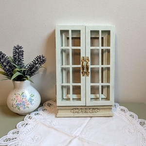 Könnte beinhalten: Ein kleiner, cremefarbener Schrank mit zwei Türen und einem Fenstergitter-Design. Der Schrank hat goldfarbene Griffe und steht auf einem dekorativen Sockel. Eine Vase mit Lavendel und eine Vase mit Blumenmuster sind ebenfalls im Bild.