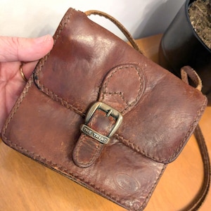 Könnte beinhalten: Kleine, braune Leder-Umhängetasche mit Klappenverschluss und silberner Schnalle. Auf der Schnalle steht "THE TREND". Die Tasche hat einen langen, dünnen Riemen und scheint Vintage zu sein.