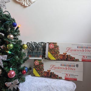 Peut inclure: Un petit sapin de Noël artificiel décoré de boules colorées et de nœuds argentés, placé à côté de deux boîtes rectangulaires de guirlandes lumineuses miniatures General Electric Lumières de Fêtes, contenant chacune 20 ampoules pour usage intérieur.