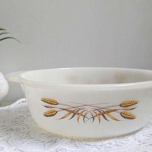 Puede incluir: Cuenco Pyrex vintage de color crema con diseño de espigas de trigo. El cuenco tiene dos asas y un borde ancho. El diseño de trigo es en tonos marrones y dorados. Un pequeño jarrón con flores está en el fondo.