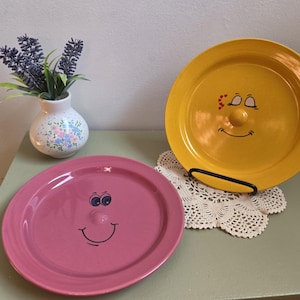 May include: Two round ceramic plates, one yellow and one pink, each featuring a raised centre nose and a hand-painted smiling face. The yellow plate displays closed eyes with red hearts, while the pink plate has blue eyes. A small vase with lavender flowers sits nearby.
