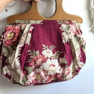 May include: A fabric handbag featuring a deep burgundy background with a floral pattern of pink, white, and green flowers and leaves. The bag has a structured, light-toned wooden handle with a circular cutout, and the fabric is pleated.