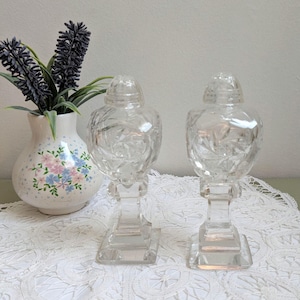 May include: Two clear, cut-glass candle holders with matching lids. The candle holders have a square base, a rectangular stem, and a bulbous top with a lid. A small vase with a floral design and lavender sprigs is in the background.