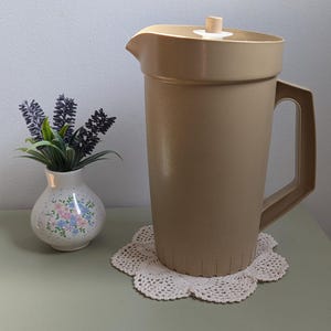Può includere: Una brocca alta in plastica beige con coperchio abbinato e manico laterale. La brocca presenta un semplice beccuccio e un design a coste verticali sulla base. È posizionata accanto a un piccolo vaso bianco con motivo floreale.