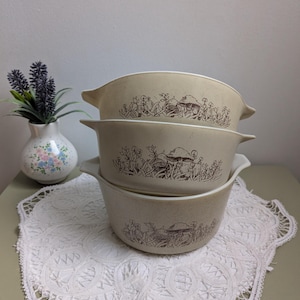 May include: Three beige casserole dishes with brown mushroom and plant designs are stacked. Each dish has a white interior and handles. They sit on a white lace doily, with a small vase of lavender and a floral vase in the background.
