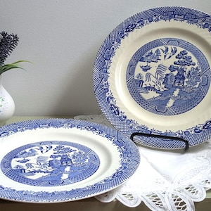 May include: Two round ceramic dinner plates featuring a blue and white willow pattern with a central landscape scene of a pagoda, bridge, and birds, surrounded by a geometric border. The plates have a cream-colored base with blue transferware.