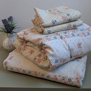 May include: A folded bedding set featuring a cream-coloured fabric with a repeating pattern of small blue and pink floral clusters and delicate dotted lines. The set includes a large comforter and matching pillowcases, all with a consistent floral motif.