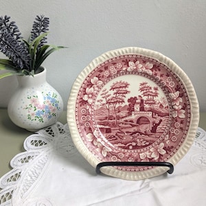 Może przedstawiać: Okrągły talerz ceramiczny z ciemnoczerwonym wzorem transferowym przedstawiającym kamienny most, drzewa i budynek. Talerz ma kremowy, falowany brzeg z motywem kwiatowym. Jest wyeksponowany na czarnym metalowym stojaku na białej koronkowej serwecie.