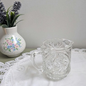 Peut inclure: Une crémière en verre taillé transparent avec une poignée et un bec verseur. La crémière présente un motif en forme d'éclat d'étoile sur le fond et les côtés. Un petit vase blanc avec un motif floral et une branche de lavande en arrière-plan.