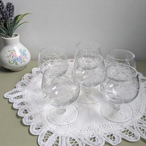 Può includere: Sei bicchieri da brandy in cristallo trasparente con disegni floreali e geometrici incisi sono disposti su una tovaglietta di pizzo bianco. Un piccolo vaso bianco con un motivo floreale e rametti di lavanda sullo sfondo.