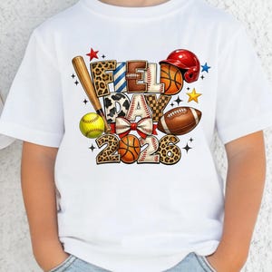 May include: Two children wearing white t-shirts with the words "Field Day 2023" in a colorful design. The design includes a baseball bat, a football, a baseball, a basketball, and a softball. The children are wearing sunglasses and blue jeans.