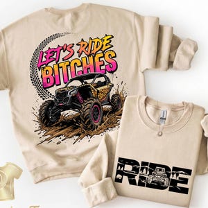 May include: Two beige sweatshirts. One features a graphic of an off-road vehicle with the text "LET'S RIDE BITCHES". The other has the word "RIDE" with an off-road vehicle graphic. Both have long sleeves.