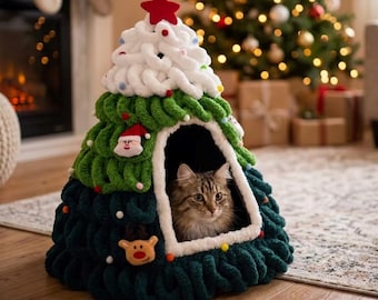 Puede incluir: Una cama para mascotas en forma de árbol de Navidad, hecha de lana tejida de color verde oscuro, con una parte superior blanca y una estrella roja. La cama tiene una abertura frontal y está decorada con pequeños adornos de fieltro de Papá Noel y un reno.
