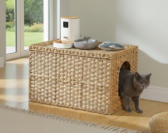 Puede incluir: Una casa para gatos rectangular de mimbre trenzado con entrada lateral arqueada, con una superficie superior plana que sostiene un comedero automático blanco y un cuenco de agua gris, junto a un juguete de pez y un gato gris saliendo.