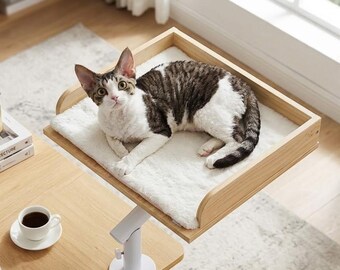 Può includere: Un gatto tigrato marrone e bianco riposa su una piattaforma rettangolare in legno con un cuscino in peluche bianco. La piattaforma è fissata a un supporto a morsetto regolabile in metallo bianco montato su una superficie in legno chiaro.