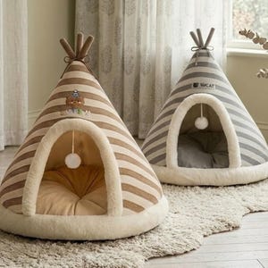 May include: Two teepee-shaped pet beds. One is striped in brown and cream, with a bear graphic. The other is striped in gray and white, with the text "LUCAT". Both have a fluffy white trim around the entrance and a hanging pom-pom.