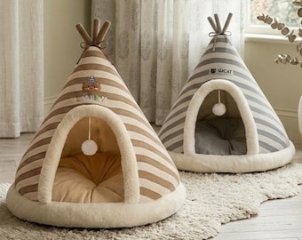 Puede incluir: Dos camas para mascotas con forma de tipi. Una está a rayas marrones y crema, con un dibujo de oso. La otra está a rayas grises y blancas, con el texto "LUCAT". Ambas tienen un borde blanco y esponjoso alrededor de la entrada y un pompón colgante.