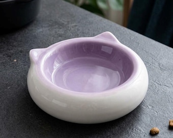 Puede incluir: Un cuenco redondo de cerámica con un exterior blanco liso y un interior de color violeta claro brillante. El borde del cuenco presenta dos pequeñas protuberancias puntiagudas que parecen orejas de gato, dando una silueta distintiva.