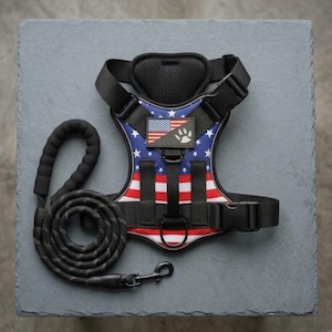 May include: A black pet harness featuring a red, white, and blue American flag pattern with a rectangular patch displaying a flag and a paw print. The harness includes black adjustable straps, a padded chest piece, and a matching black rope leash.