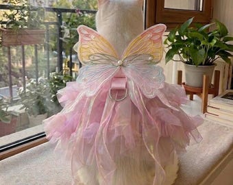 Puede incluir: Un gato blanco con un vestido de hada rosa e iridiscente con alas de mariposa a juego. El vestido tiene capas de tela con volantes y un arnés rosa con un anillo plateado. Las alas son de colores pastel con detalles de perlas.