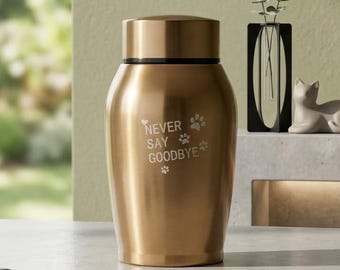 Puede incluir: Una urna de metal dorado con tapa cilíndrica y cuerpo liso y cónico. La parte frontal presenta el texto grabado NEVER SAY GOODBYE, acompañado de pequeños iconos de corazones y huellas de patas. El recipiente tiene un acabado pulido.