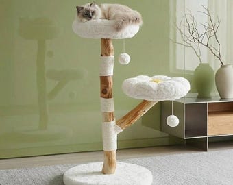 Puede incluir: Un árbol para gatos con un tronco de madera natural y tres secciones envueltas en cuerda de sisal color crema. Tiene dos plataformas blancas de felpa con forma de flor y una base redonda. Un gato esponjoso descansa arriba con dos pompones colgantes.