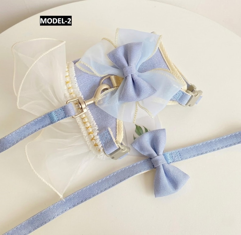 Puede incluir: Un arn&eacute;s para mascota de color azul claro con correa a juego etiquetado como MODEL-2. El arn&eacute;s presenta un gran lazo de organza transparente, una hilera de perlas blancas de imitaci&oacute;n y una anilla en D de metal. La correa incluye un lazo peque&ntilde;o.