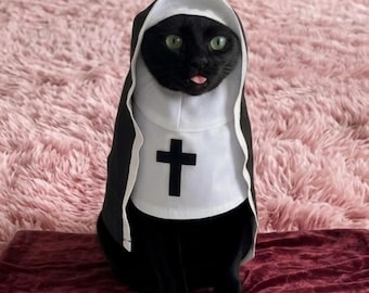 Puede incluir: Un gato negro con un disfraz de monja blanco y negro. El disfraz cuenta con una toca blanca y un panel frontal blanco con un símbolo de cruz negra centrado. El gato tiene ojos verdes y se le puede ver una pequeña parte de la lengua.