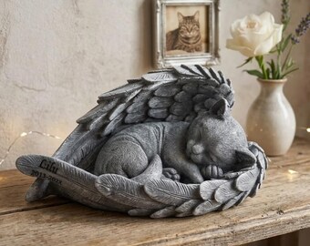 Puede incluir: Una figura de gato de efecto piedra gris durmiendo acurrucada dentro de un par de grandes alas emplumadas. El ala presenta el texto grabado Lilu y 2013-202X. La escultura tiene un acabado texturizado y representa a un gato descansando.