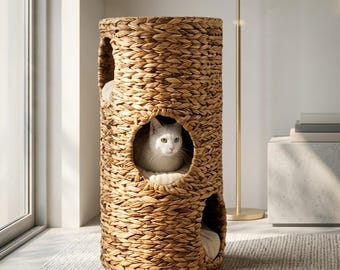 Puede incluir: Una torre cilíndrica para gatos hecha de fibras naturales tejidas con tres aberturas circulares. Un gato blanco descansa dentro de la abertura central sobre un cojín suave y claro. La estructura tiene una apariencia trenzada y texturizada.