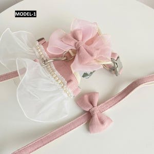Puede incluir: Un arn&eacute;s para mascota de color rosa con correa a juego etiquetado como MODEL-1. El arn&eacute;s presenta un gran lazo de organza rosa, ribete de volantes blancos y una hilera de peque&ntilde;as perlas blancas. La correa incluye un peque&ntilde;o lazo de tela.