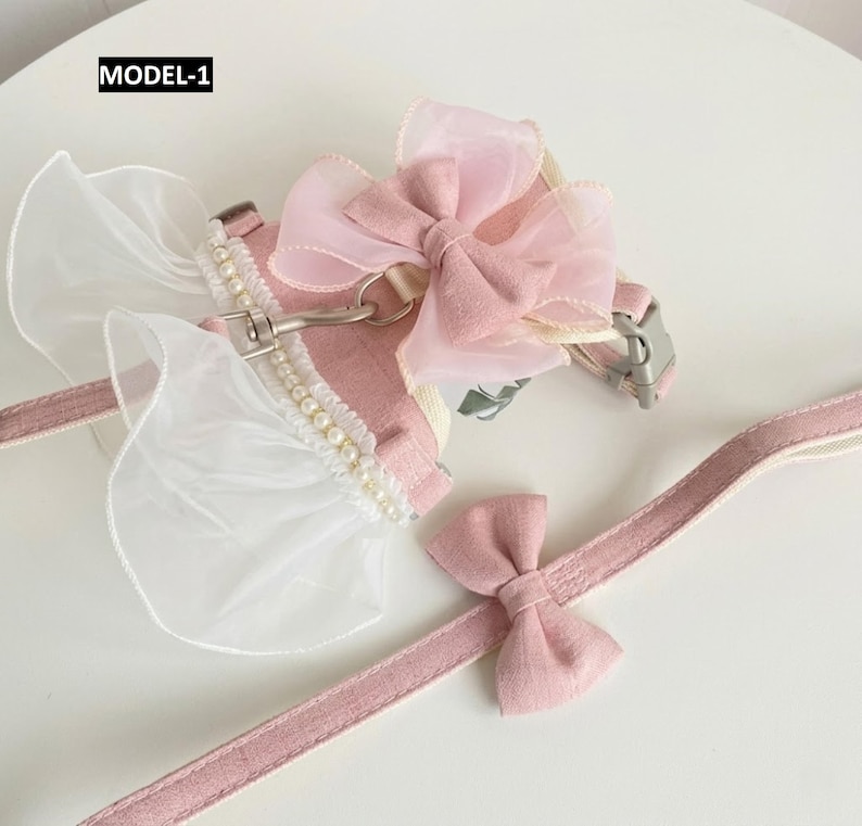Puede incluir: Un arn&eacute;s para mascota de color rosa con correa a juego etiquetado como MODEL-1. El arn&eacute;s presenta un gran lazo de organza rosa, ribete de volantes blancos y una hilera de peque&ntilde;as perlas blancas. La correa incluye un peque&ntilde;o lazo de tela.