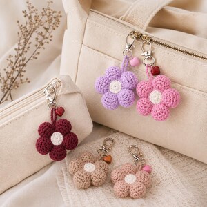 Könnte beinhalten: Gehäkelte Blumen-Schlüsselanhänger in Lila, Rosa, Burgunderrot und Beige. Jede Blume hat eine weiße Mitte und einen kleinen Herzanhänger. Die Schlüsselanhänger sind an silberfarbenen Metallverschlüssen befestigt.