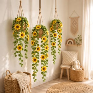 May include: Three hanging crochet sunflower baskets with yellow flowers and green leaves. The baskets are suspended by brown rope and are against a white wall. A woven basket and rug are in the foreground.