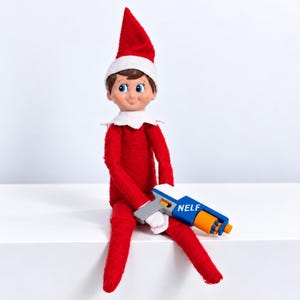 May include: A plush elf doll wearing a red suit with a white collar and a matching red pointed hat. The doll has brown hair, blue eyes, and is holding a small blue, white, and orange toy blaster with the word NELF printed on the side.