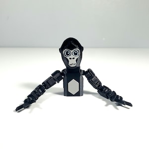 Puede incluir: Una figura de gorila articulada de color negro con cara blanca, ojos grandes y una placa pectoral hexagonal blanca. La figura presenta un torso segmentado y brazos largos y flexibles con múltiples articulaciones de material negro mate.