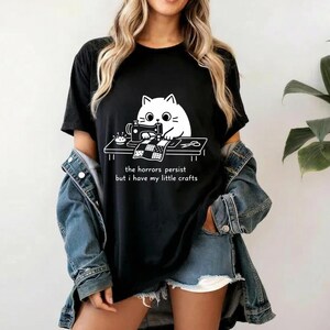 Pode incluir: Camiseta preta com um gráfico branco de um gato costurando em uma mesa. O gráfico inclui uma máquina de costura e uma pequena mesa. O texto "the horrors persist but i have my little crafts" está abaixo. Roupa casual.