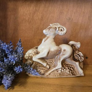 May include: A ceramic figurine of a ram in a reclining pose, with a cream-colored body and brown accents. The ram rests on a textured base with floral details. A bundle of blue flowers is to the left of the figurine.