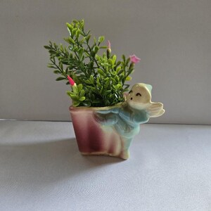 May include: A ceramic planter featuring a sculpted figure with wings attached to the side. The planter has a gradient finish transitioning from a deep reddish-pink at the base to a pale blue-green on the figure. It contains a small artificial plant.