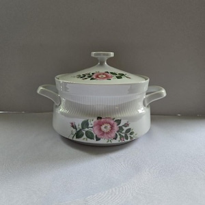 May include: A white ceramic lidded tureen featuring a delicate pink rose floral pattern with green leaves on the body and lid. The vessel has two side handles and a central knob on the lid, with a subtle vertical ribbed texture around the upper rim.