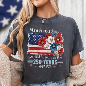 Può includere: Una t-shirt grigio antracite a maniche corte con una grafica della bandiera americana decorata con fiori rossi, bianchi e blu. Il testo recita America, God shed his grace on thee, 250 YEARS e SINCE 1776, con piccole stelle rosse.