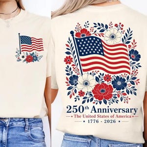 Puede incluir: Una camiseta color crema con una pequeña bandera estadounidense y un diseño floral en el pecho izquierdo. La parte trasera muestra una gran bandera estadounidense rodeada de flores rojas, blancas y azules con el texto 250th Anniversary The United States of America 1776-2026.