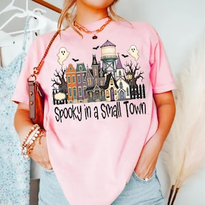 Op de afbeelding: Een lichtroze t-shirt met een afbeelding van een klein stadje met kleurrijke huizen, een watertoren, kale bomen en twee geesten. De tekst "Spooky in a Small Town" staat in het zwart onder de illustratie. Het shirt heeft een ronde hals.