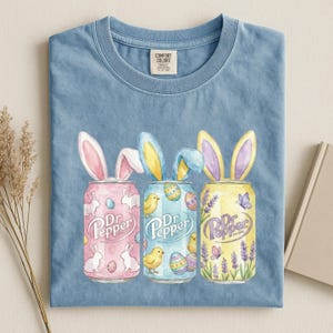May include: A light blue t-shirt featuring three Dr. Pepper soda cans with bunny ears. The cans are pink, blue, and yellow, decorated with Easter-themed graphics. The shirt has a Comfort Colors tag.