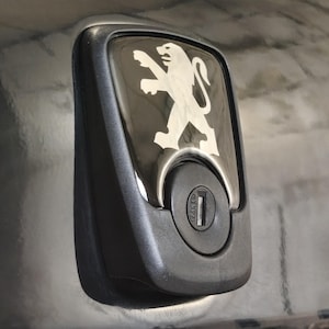 Peut inclure: Gros plan d'une trappe à carburant de voiture noire avec un emblème chromé. L'emblème représente un lion blanc, le logo Peugeot. La trappe à carburant a une serrure et un contour en plastique noir.