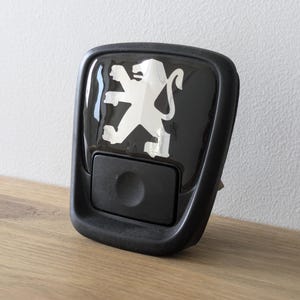 Op de afbeelding: Zwart rechthoekig auto-onderdeel met een glanzende afwerking. Het heeft een wit Peugeot-logo en een kleine, zwarte, ronde knop eronder. Het onderdeel staat tegen een lichte muur en een houten oppervlak.