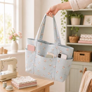 May include: A light blue tote bag featuring a repeating pattern of small pink flowers and green leaves. The bag has two shoulder straps and three exterior pockets on the front, which contain a small notebook, a card holder, and a smartphone.