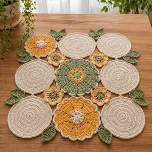 May include: Handmade crochet table runner featuring a floral design. The runner is made of cream, yellow, and green yarn, with circular and flower-shaped motifs. The table runner is approximately 24 inches square.